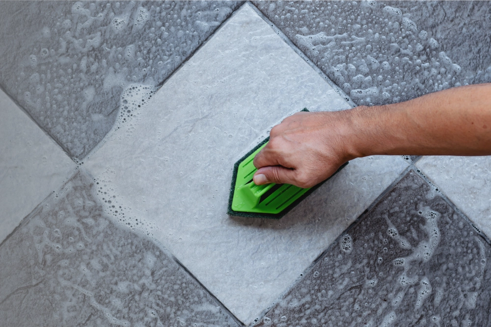 Close-up of a hand using a scrub brush and cleaning solution on gray floor tiles, demonstrating how to clean tile floors and remove dirt